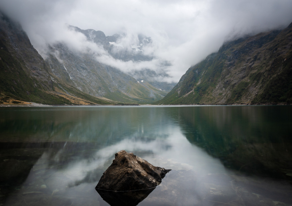 14 Incredible Hikes On the South Island of New Zealand 11 A lake with a rock in the foreground at mountains behind, partially concealed by clouds at Lake Marian.