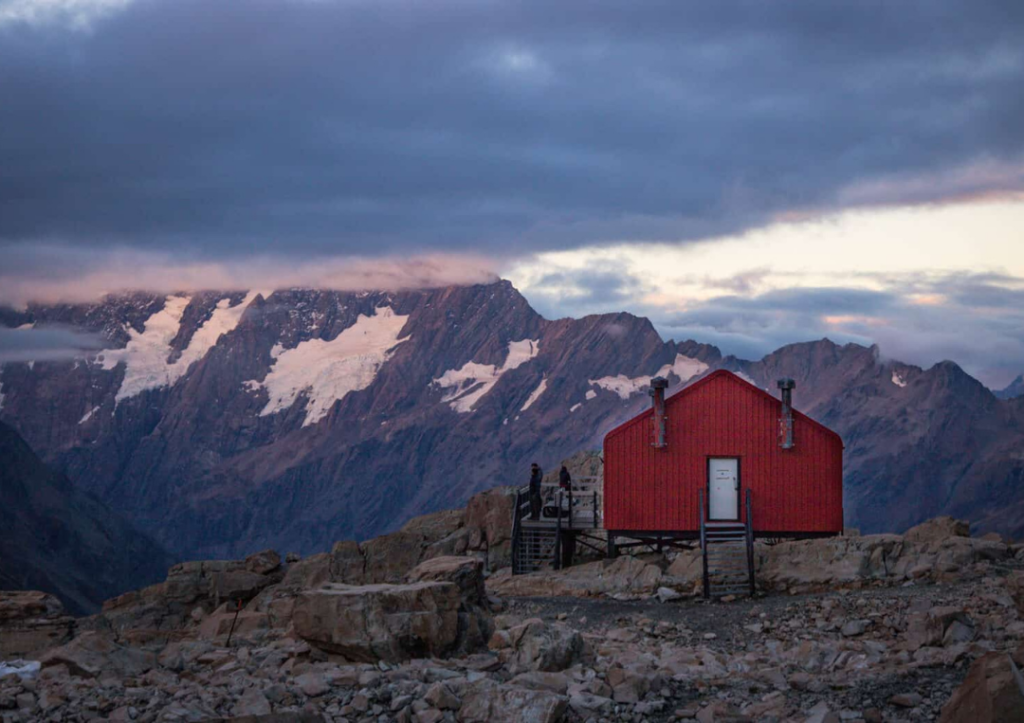 14 Incredible Hikes On the South Island of New Zealand 7 A red hut at sunrise with mountains pink in the sunlight behind.
