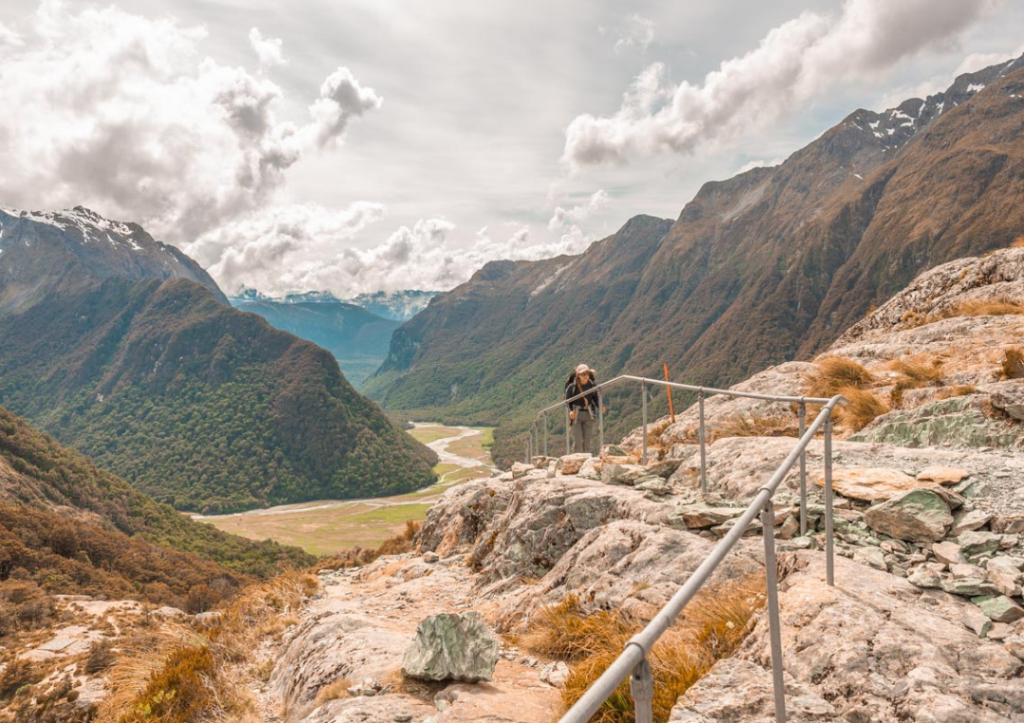 14 Incredible Hikes On the South Island of New Zealand 6 Person walking up some steps on a rocky trail with mountains behind on the Routeburn Track.