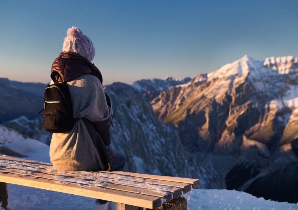 Woman in a coat and hat sitting on a bench in the snowy mountains. 