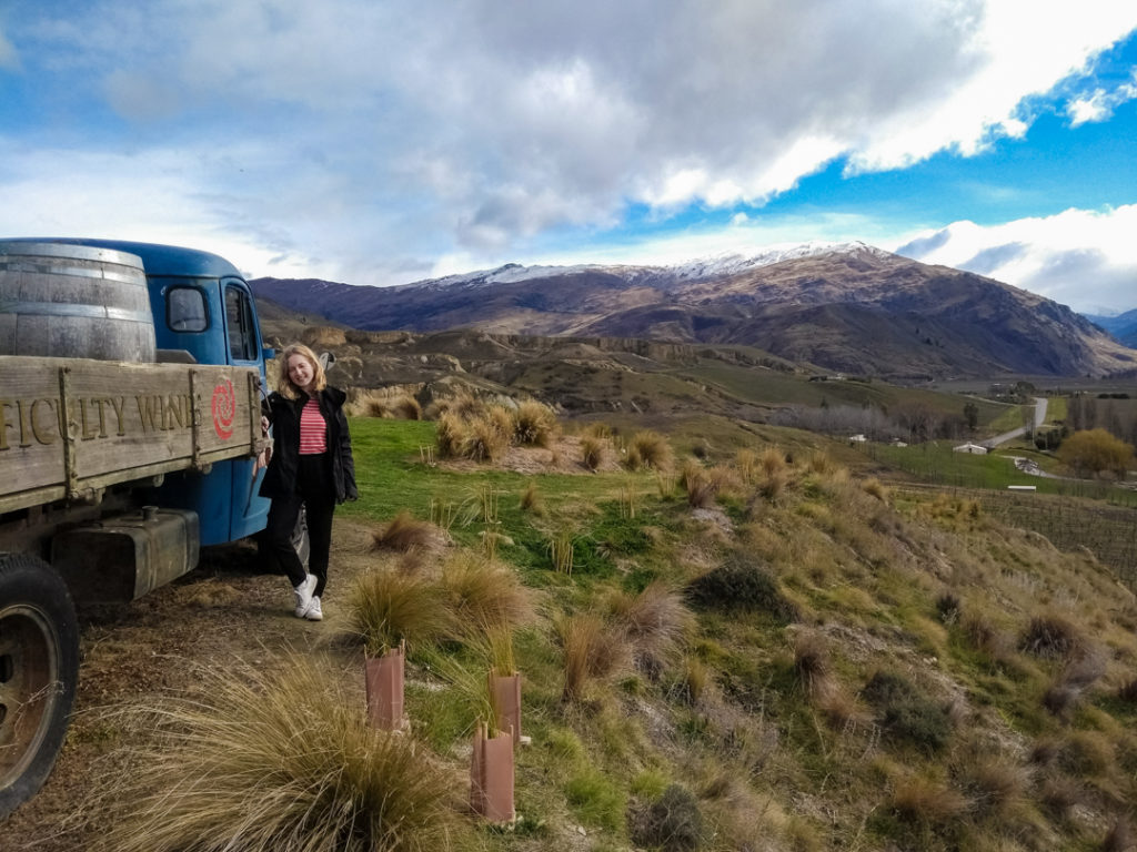 Girl standing by a retro truck in the hills with snow-capped mountains behind on this New Zealand packing list. 