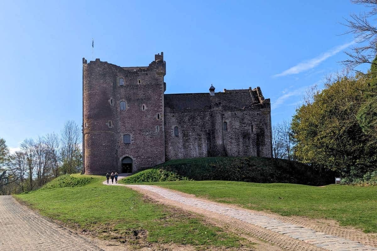 An almost intact fortress castle stands on a grassy mound in some shadow. Below them are three people walking along the path up to it. This is Doune Castle in Scotland on the Outlander tour. 