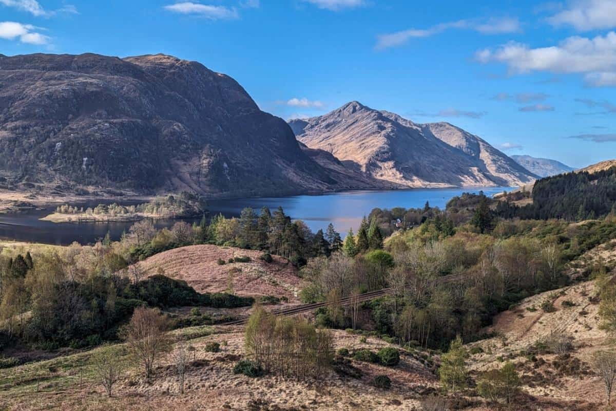 Best 10-day Scottish Highlands itinerary (No Car Needed!) 2 A tranquil loch winds through a valley surrounded by rugged, sunlit mountains and forested slopes under a bright blue sky.