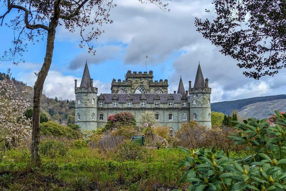 Inveraray Castle in Scotland, a stately green-stone building with turrets and battlements, framed by blooming spring trees and lush greenery under a partly cloudy sky. A top destination on the best Scotland day tours, this castle sits nestled in the scenic hills of Argyll.