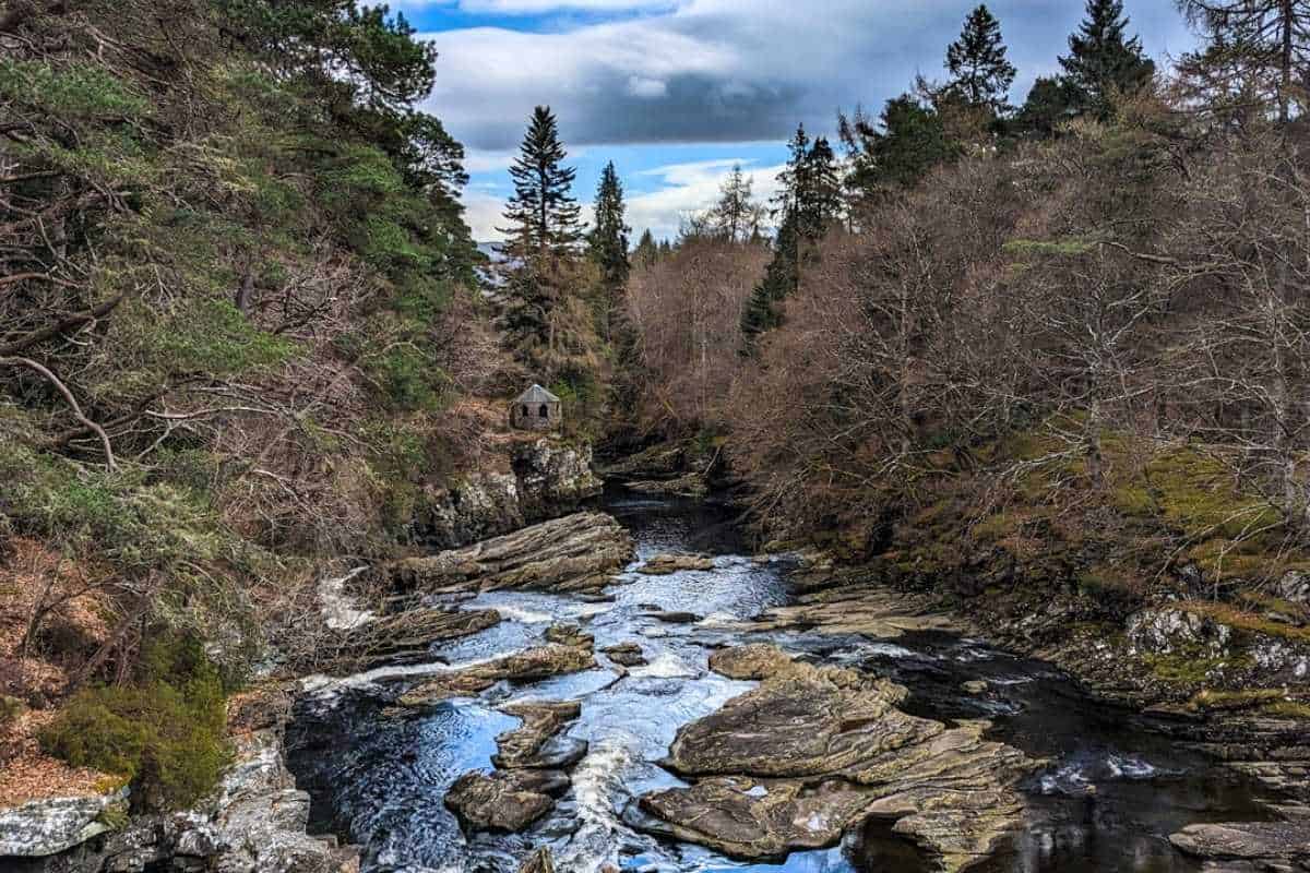 Best 10-day Scottish Highlands itinerary (No Car Needed!) 13 A rocky river cuts through a forest with tall evergreens and leafless trees, with a small stone hut perched on a cliff above the water.