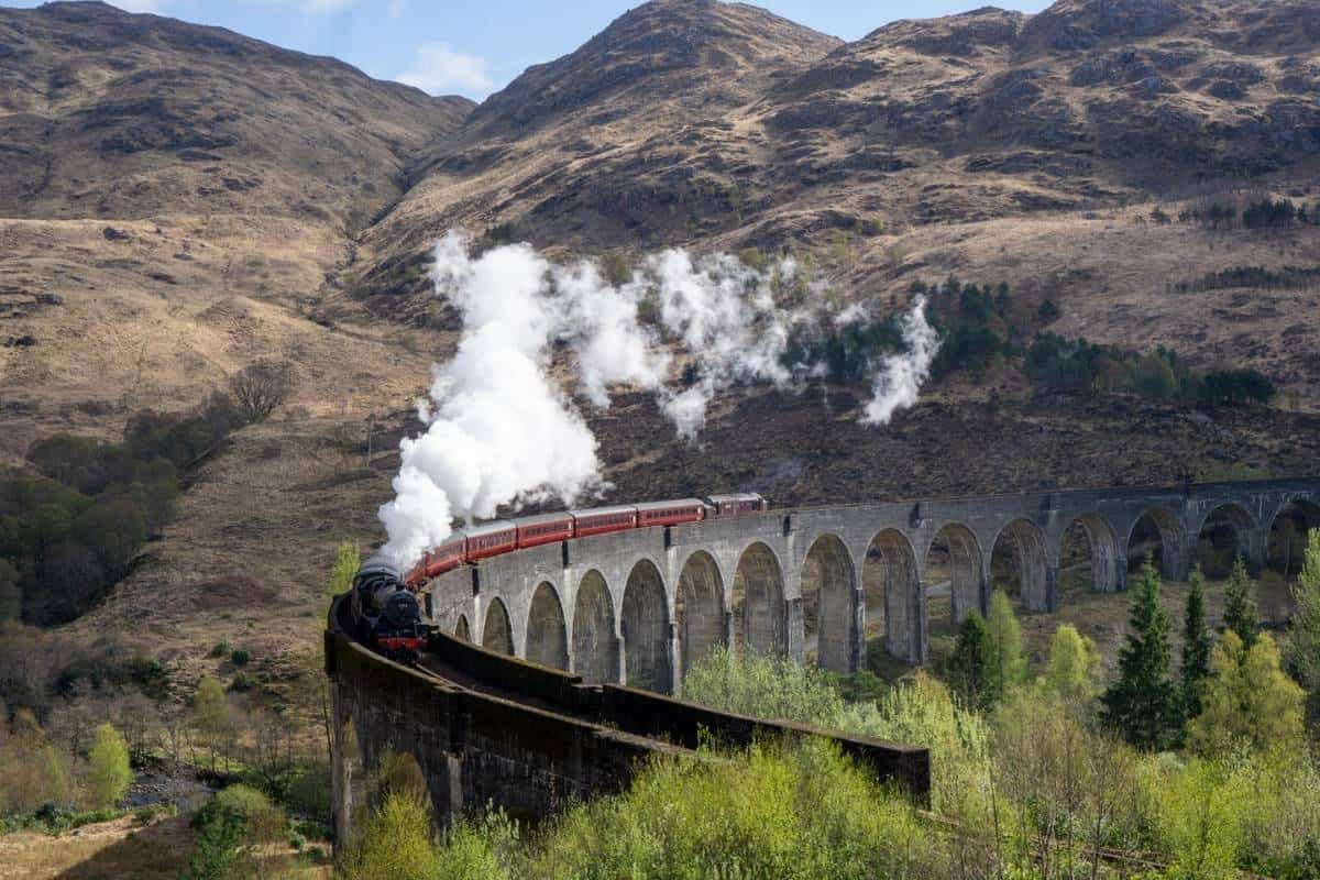 Best 10-day Scottish Highlands itinerary (No Car Needed!) 4 A black steam train with red carriages crosses the iconic Glenfinnan Viaduct, trailing white smoke against the backdrop of rolling hills. This scenic rail journey is a favorite highlight on this Scottish Highlands itinerary.