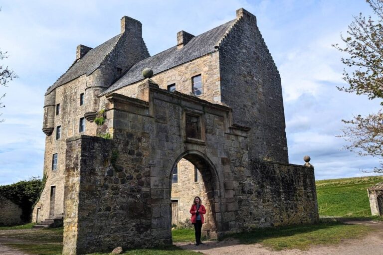 A woman wearin a red jacket stands under a stone archway facing the camera. Behind her is the tall fortress style building of Midhope Castle in Scotland.