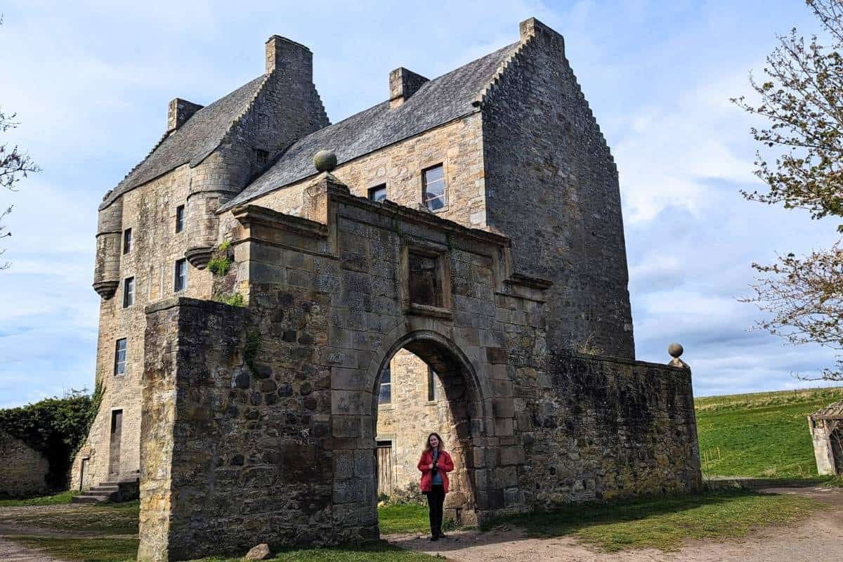 A woman wearin a red jacket stands under a stone archway facing the camera. Behind her is the tall fortress style building of Midhope Castle in Scotland. 