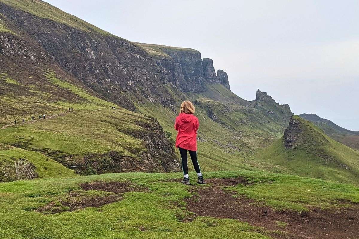 A girl with short blonde hair and a red coat stands with her back to the camera. She's on a grassy ledge looking across to the dramatic Quiraing on the Isle of Skye. 