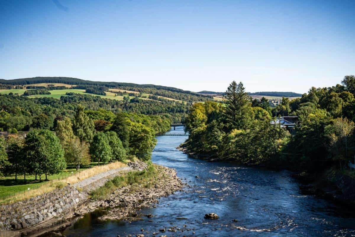 Best 10-day Scottish Highlands itinerary (No Car Needed!) 20 A winding river cuts through a lush green landscape of rolling hills and mixed forest, with a distant bridge visible beneath a cloudless blue sky.