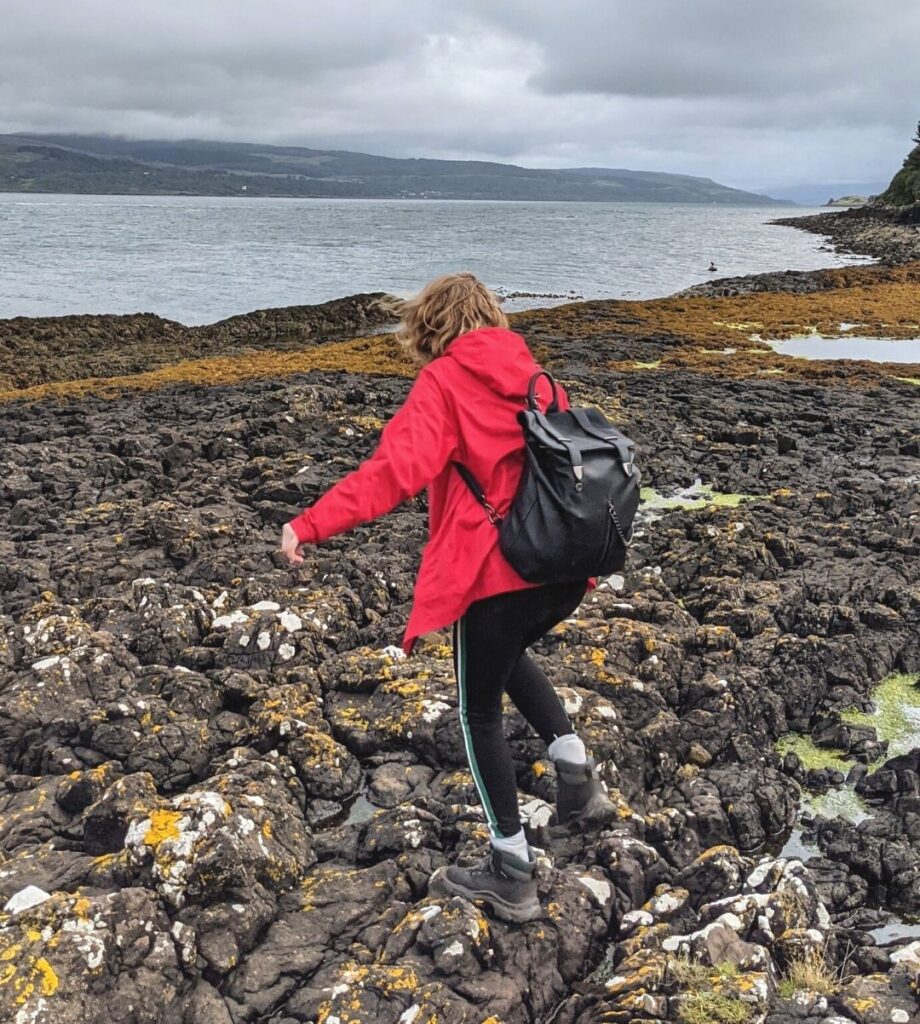 Girl wearing a red jacket, shoes, bag and leggings jumping over the rocks on a Scottish coastline. 