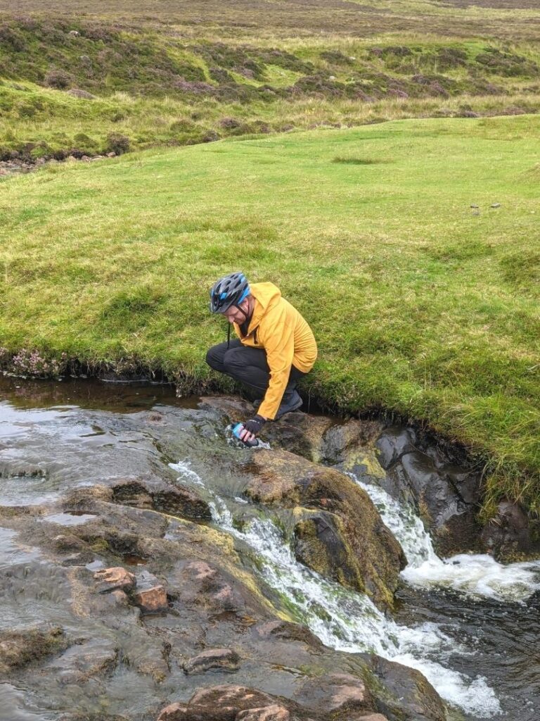 Man in a yellow jacket ad wearing a bike helmet holding a water bottle over the rocks in a brook in Scotland. 