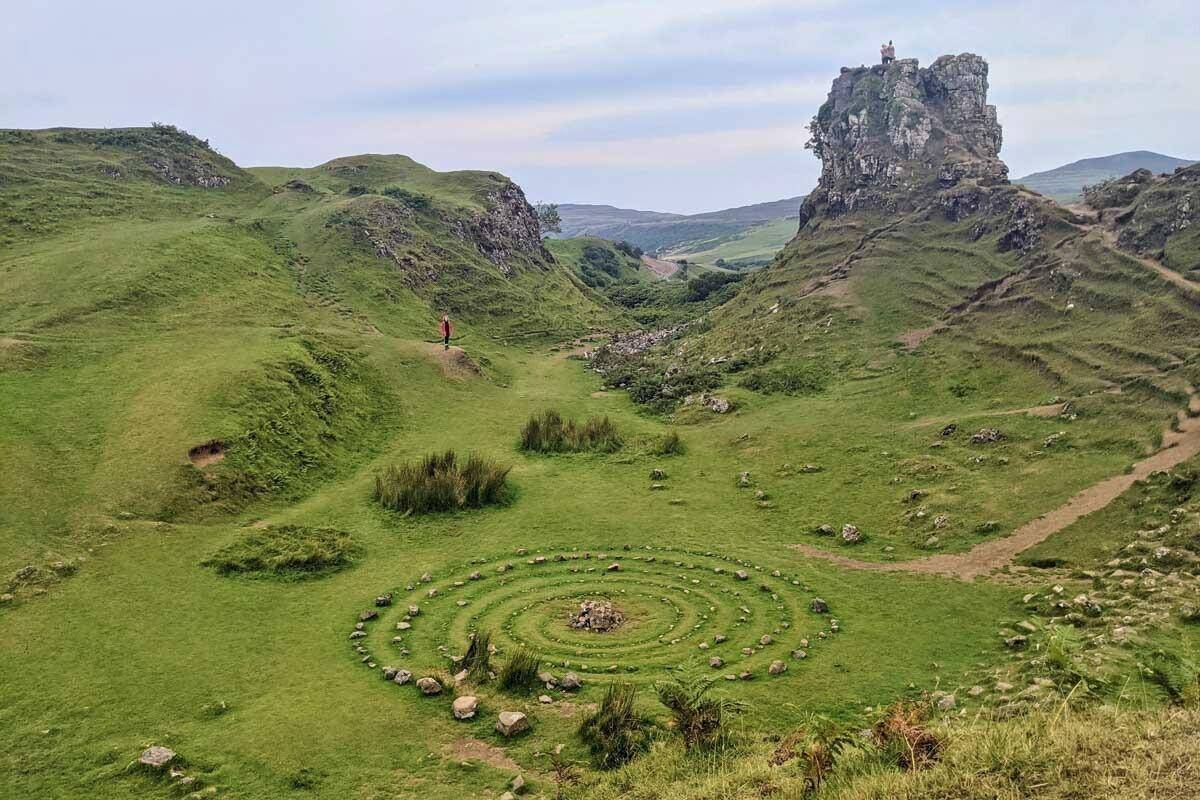 A wide view of the Fairy Glen on the Isle of Skye, featuring a striking spiral pattern of stones on green grass and a rocky hill with a person standing on top. The whimsical landscape looks like something out of a fantasy novel.