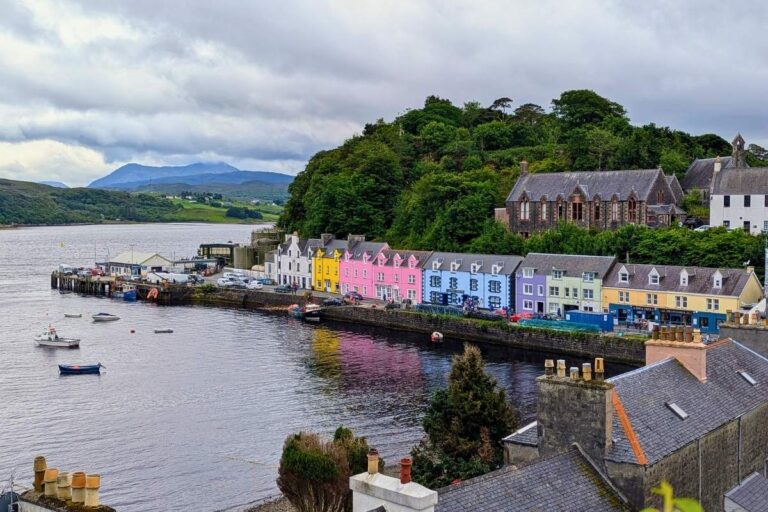 A row of brightly colored buildings lines the harbor in Portree, the main town on the Isle of Skye, with calm water reflecting the pastel facades and moored boats floating nearby. Rolling hills and a dramatic sky frame this charming waterfront scene, often included in a 2-day Isle of Skye itinerary.