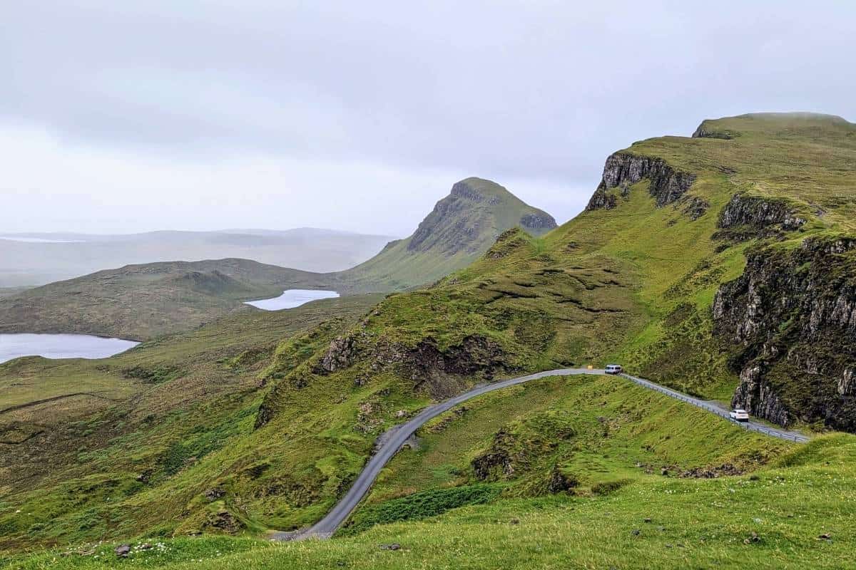A winding road cutting through the lush, rolling hills of the Quiraing on the Isle of Skye, with misty lochs visible in the distance. A perfect stop on a 2-day Isle of Skye itinerary.