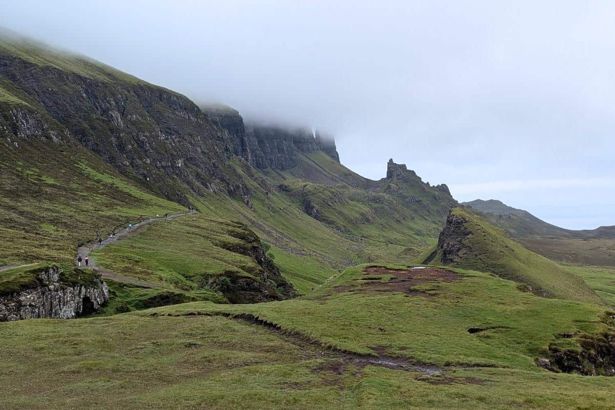 Hikers walking along a trail under a misty ridge in the Quiraing, with steep cliffs disappearing into low clouds and a rugged green landscape stretching into the distance.