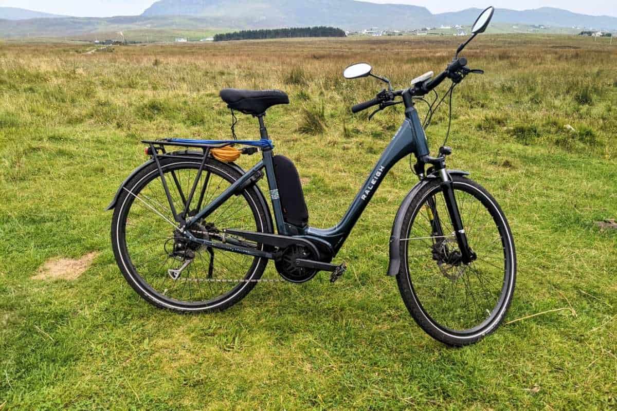 A dark teal Raleigh electric bike stands on grassy terrain with scenic highland hills in the background. Ideal eco-friendly transport for a 2-day Isle of Skye itinerary.