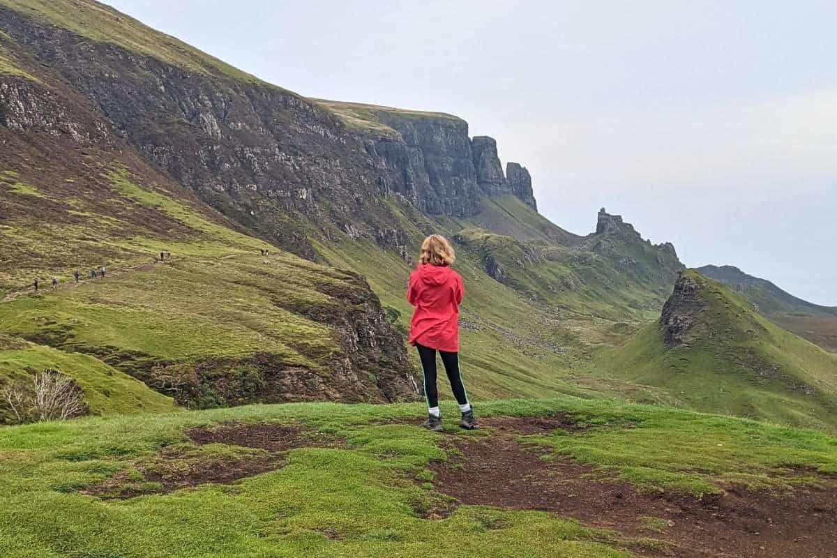 A woman in a red jacket stands on a grassy hill in the Quiraing, looking toward the dramatic jagged cliffs and spires partially covered in fog.