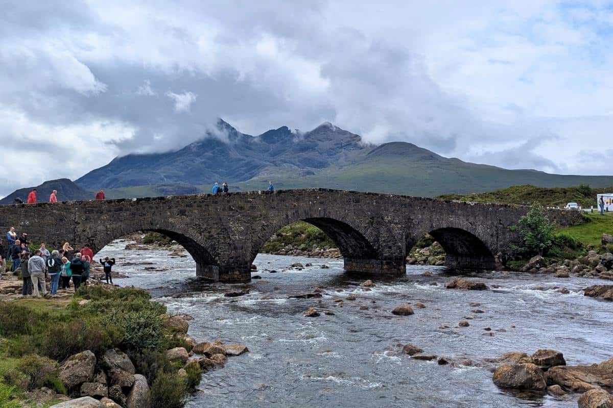 The historic Sligachan Bridge arches over a rocky stream, with tourists gathered below and misty Cuillin mountains looming in the background. A must-see landmark during a 2-day Isle of Skye itinerary.