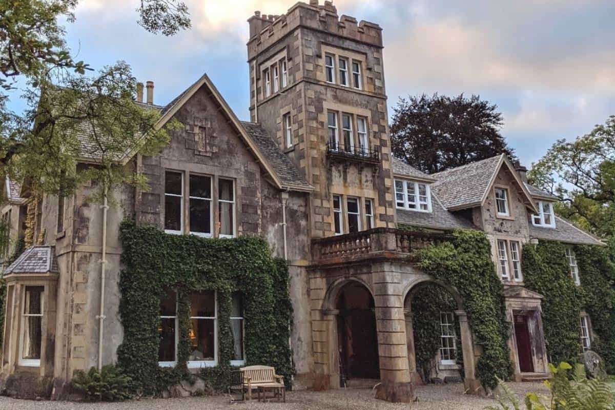 A grand ivy-covered manor house with stone towers and arched entryways, surrounded by trees and evening light casting a warm glow through the windows.