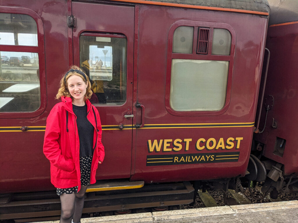 Girl in a red coat standing by a carriage at the platform for Jacobite steam train review. 