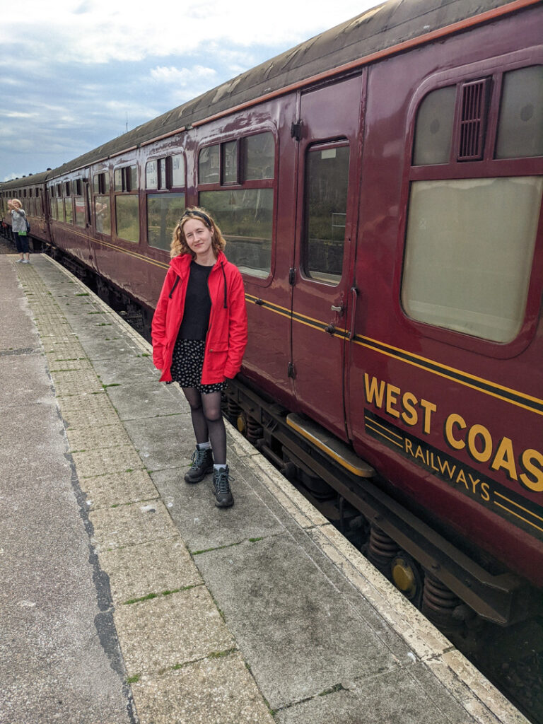 Girl in a red coat standing by a red train carriage at the platform. 