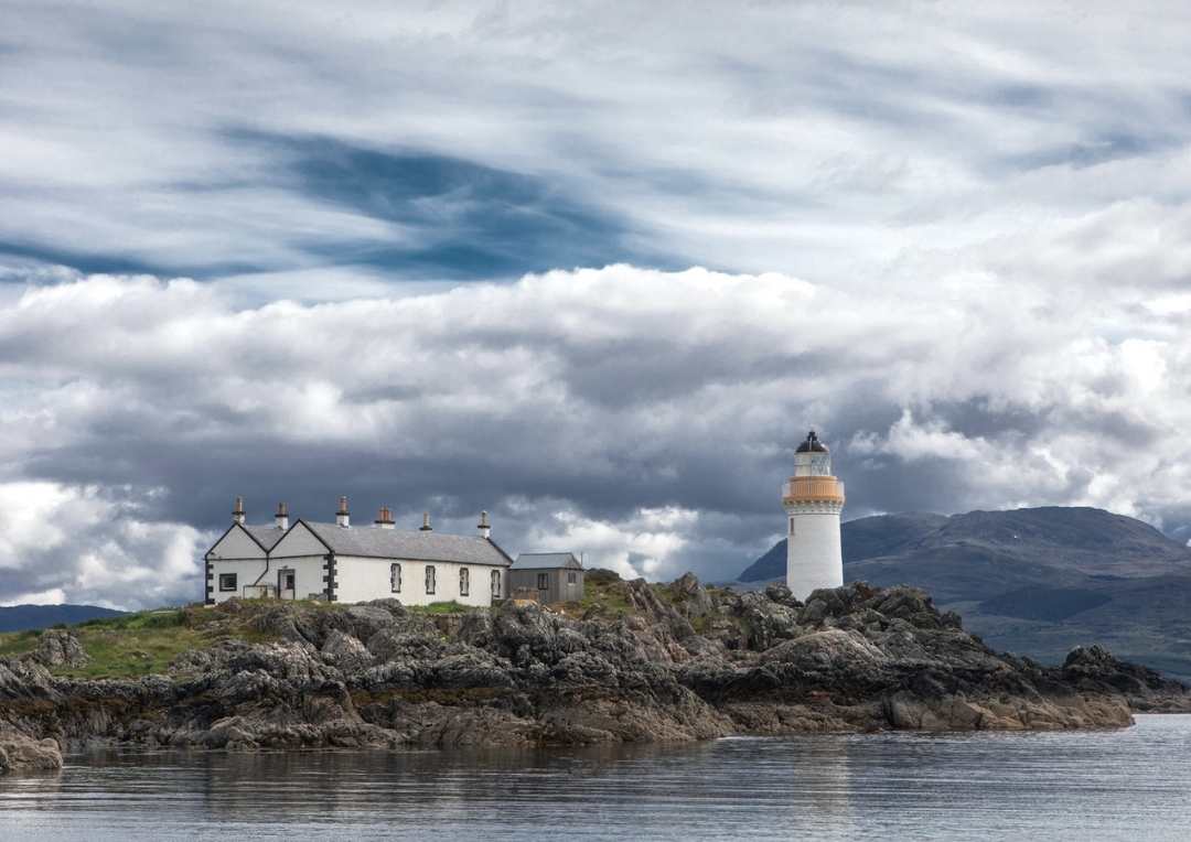 A white house with a lighthouse sitting on a rocky island and hills behind. 