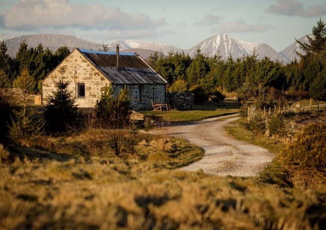 A traditional stone bothy with mountains and trees behind. Lusa Bothy is one of the best unique places to stay on Skye. 
