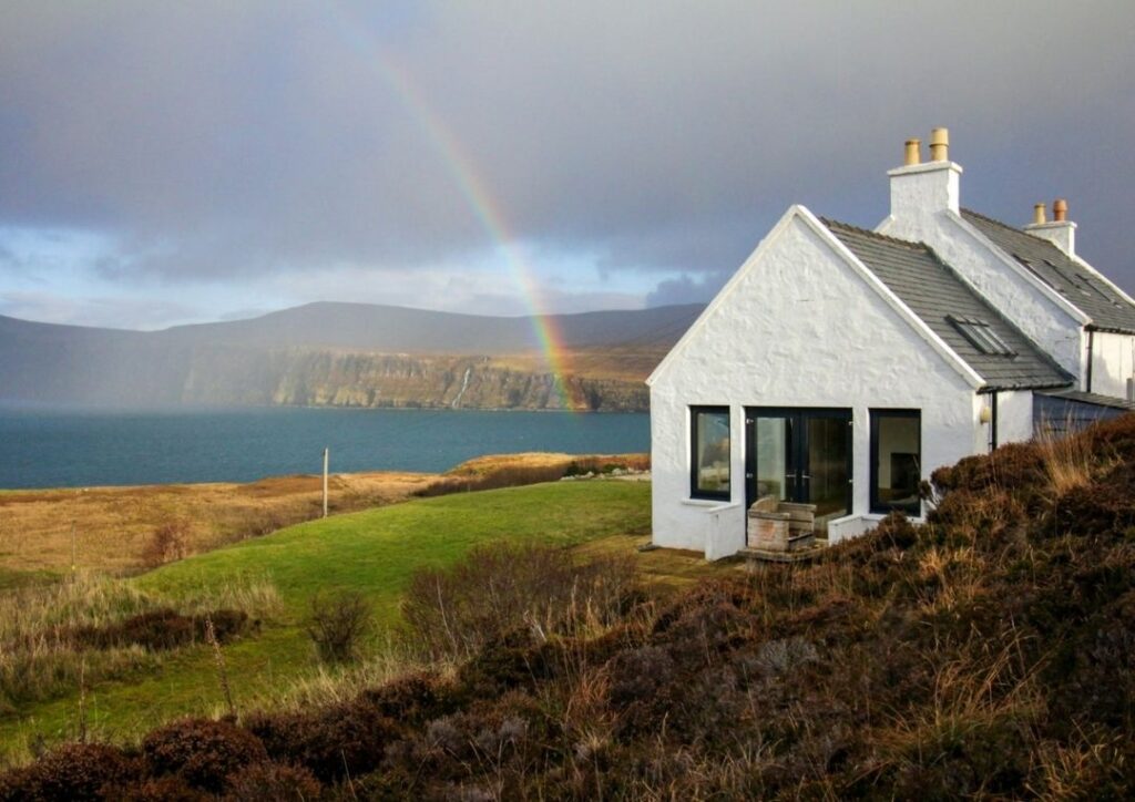 A white house on the coastline with a rainbow over the sea behind. 