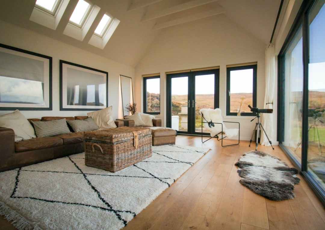 A large lounge area with a brown sofa and a white rug and large windows. Milovaig House, Skye. 