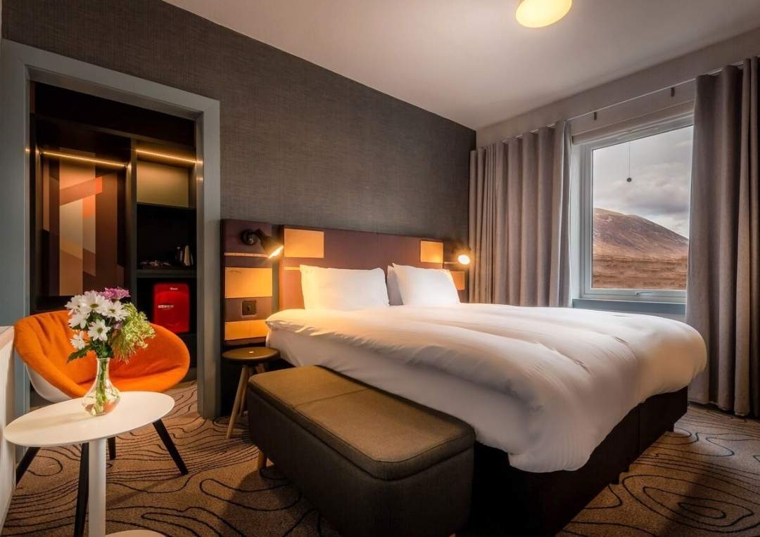A large white bed with an orange seat next to it and grey wallpaper in a room at the Sligachan Hotel. 