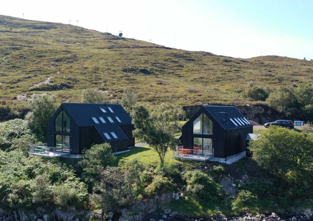 Two black huts with big windows surrounded by green hills. The Tin Sheds are one of the best places to stay on Skye. 