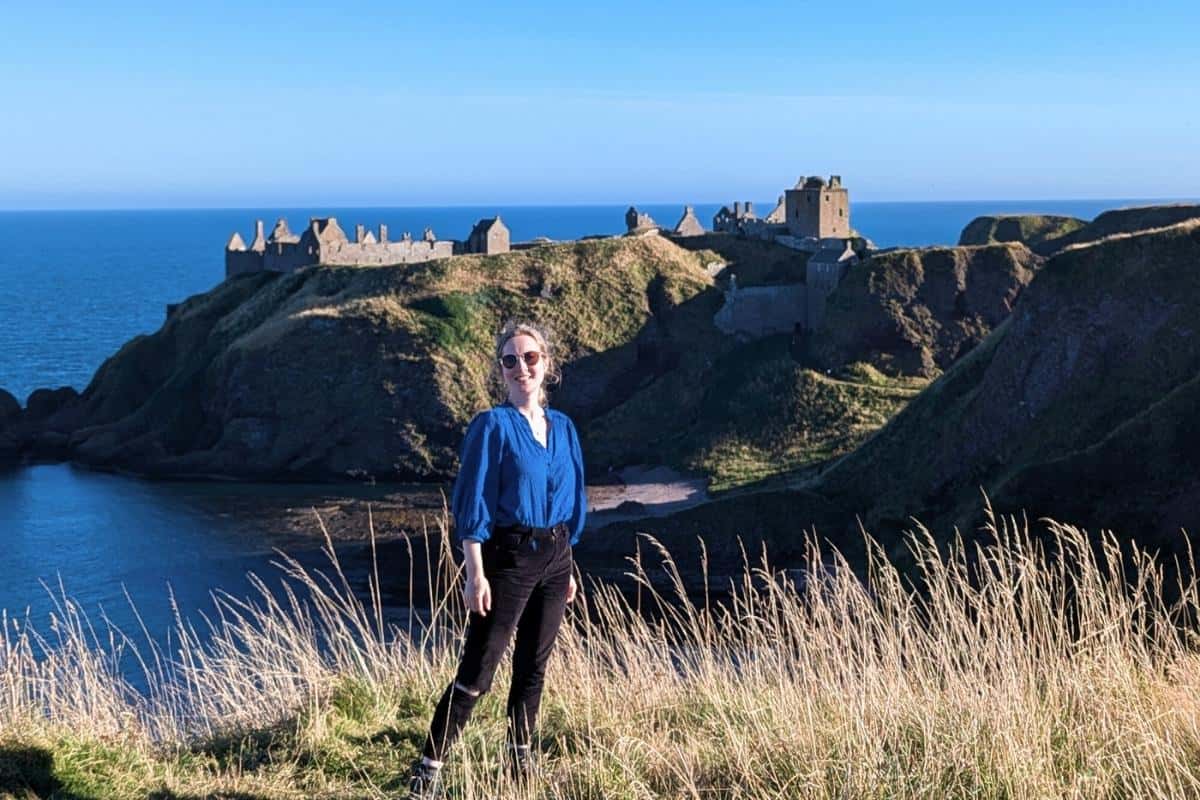 Castle ruins on a hill with the sea behind at Stonehaven in Scotland. A girl in a blue top and sunglasses smiles at the camera. 