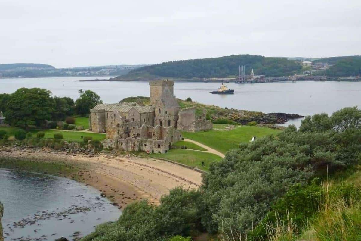 A ruined abbey sits in front of a beach on a small grassy island covered in trees. There is a small boat and the mainland in the distance. 