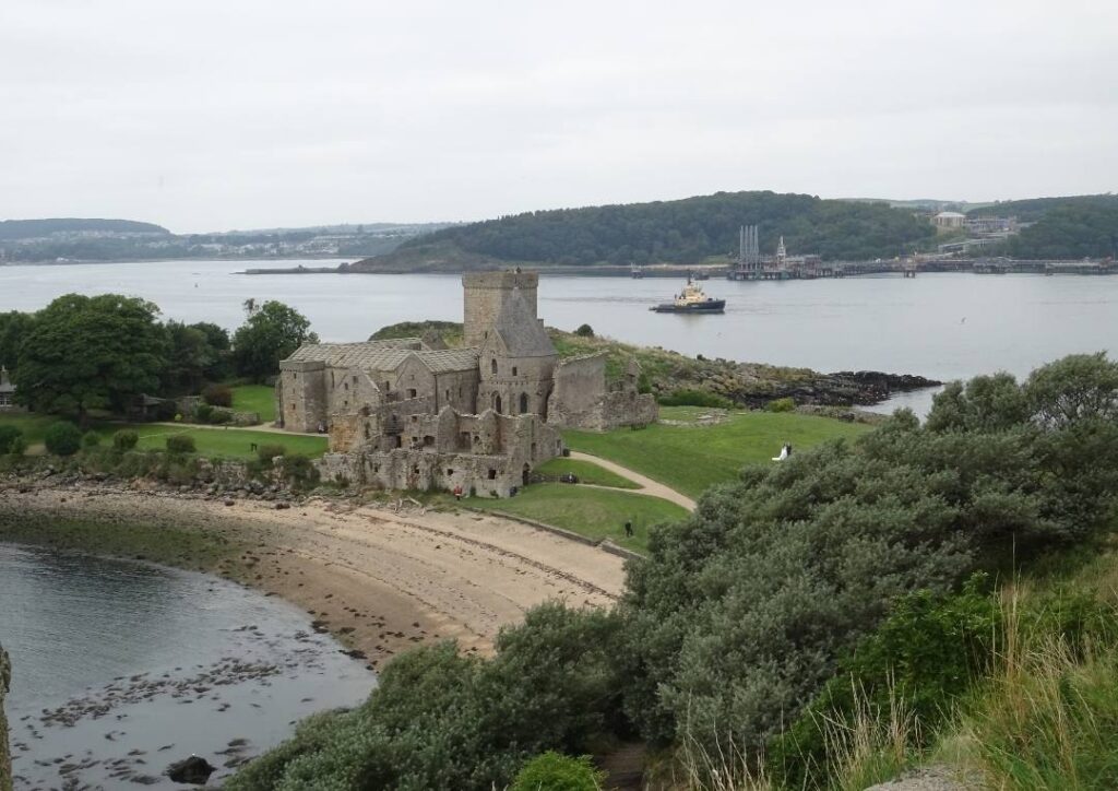 Partial ruins of an abbey on a small island and next to a beach. Edinburgh in the distance. Inchcolm Abbey is one of the best day trips from Edinburgh by ferry. 
