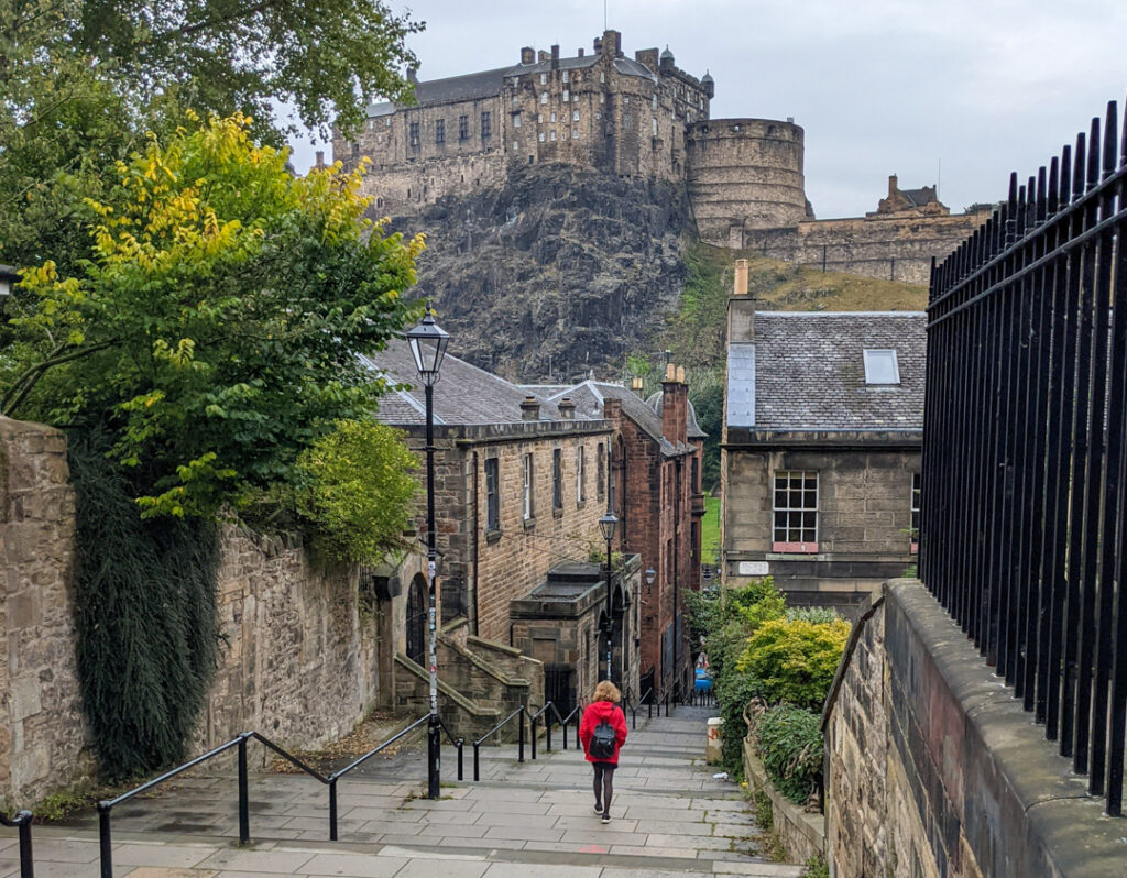 A girl in a red jacket walking down a street in Edinburgh Castle - the Vennel Steps - with Edinburgh Castle behind. 