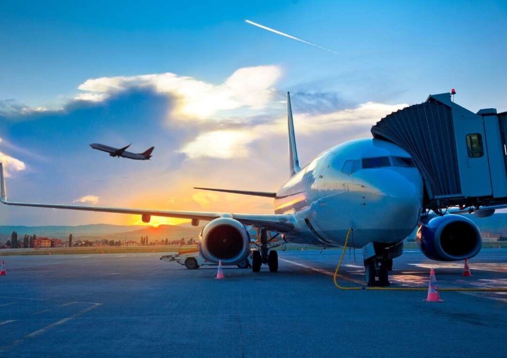 An airplane at an airport with a plane in the air behind it against the sunrise. 