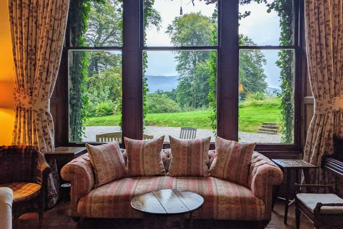 A room with large windows showing a lush garden, framed by thick curtains. There's an antique sofa in front of it with mahogany furniture. The lounge at Viewfield House. 