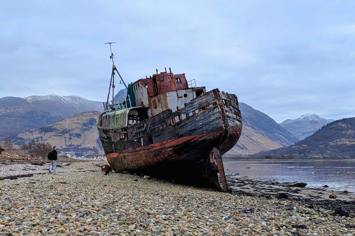 A rusting, abandoned fishing boat tilts dramatically on a rocky shore, backed by misty Scottish mountains and a cloudy sky. A lone traveler walks along the pebbled beach, adding scale to the haunting scene.