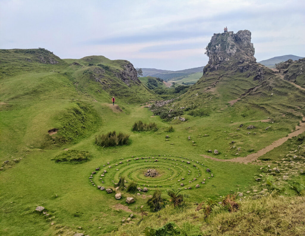 Photo of a stone spiral surrounded by small hillocks and terraces. A girl in a red coat stands in the distance. Fairy Glen on Isle of Skye. 