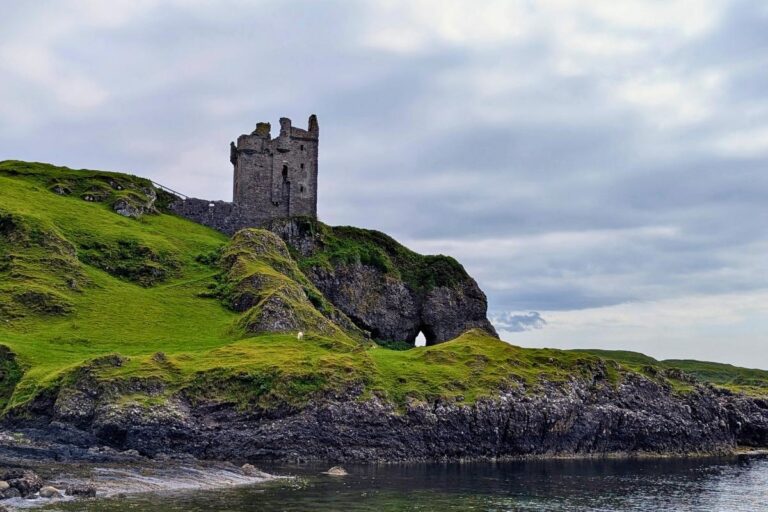 A weathered stone castle stands on a grassy cliff above the sea, with rugged rock formations and a natural arch below. The overcast sky adds a moody, historic feel to this coastal scene, typical of a Scotland west coast itinerary.