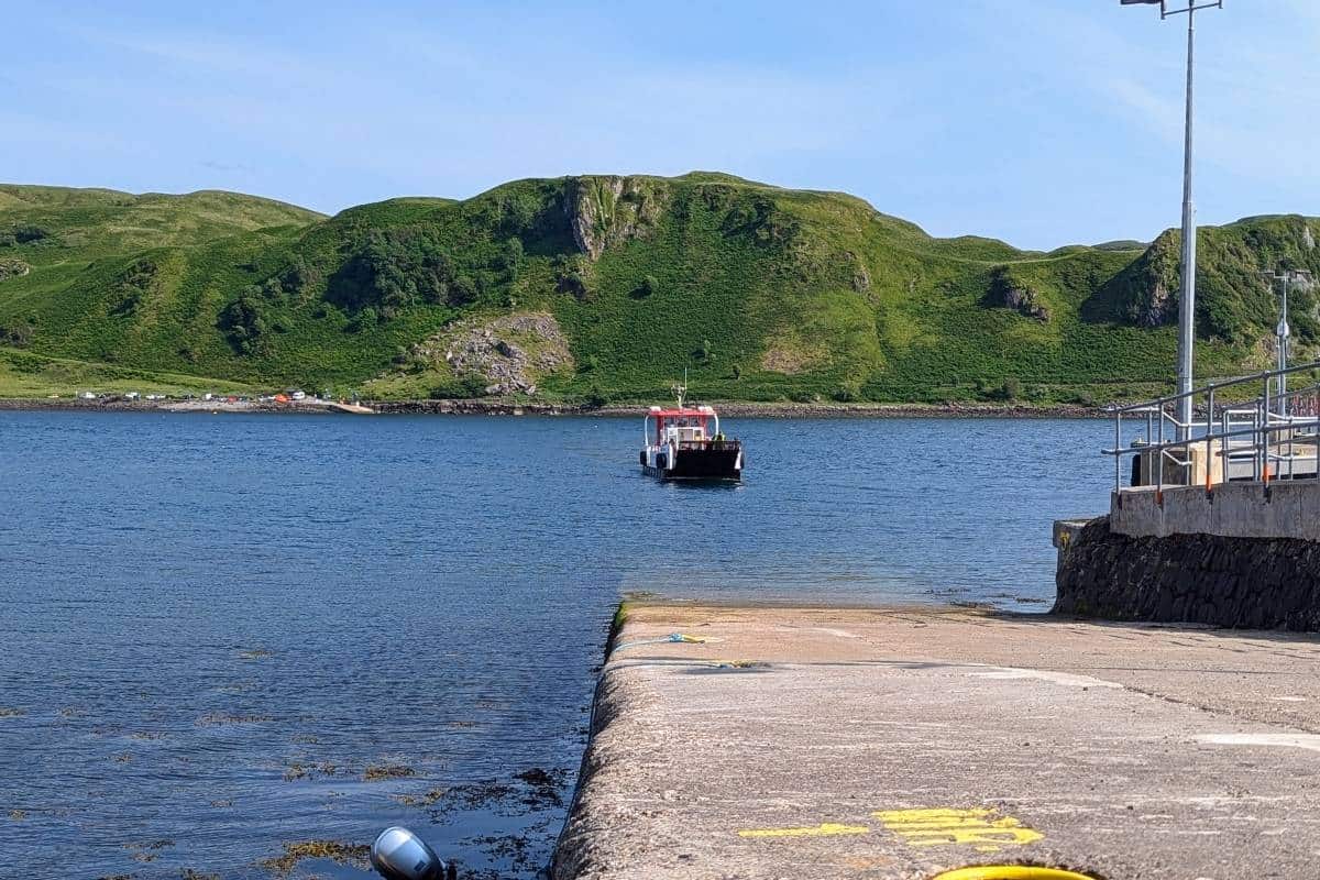 A small red-and-white ferry approaches a concrete pier, with grassy hills and scattered tents visible across the blue water. Calm weather and clear skies frame this peaceful coastal setting.