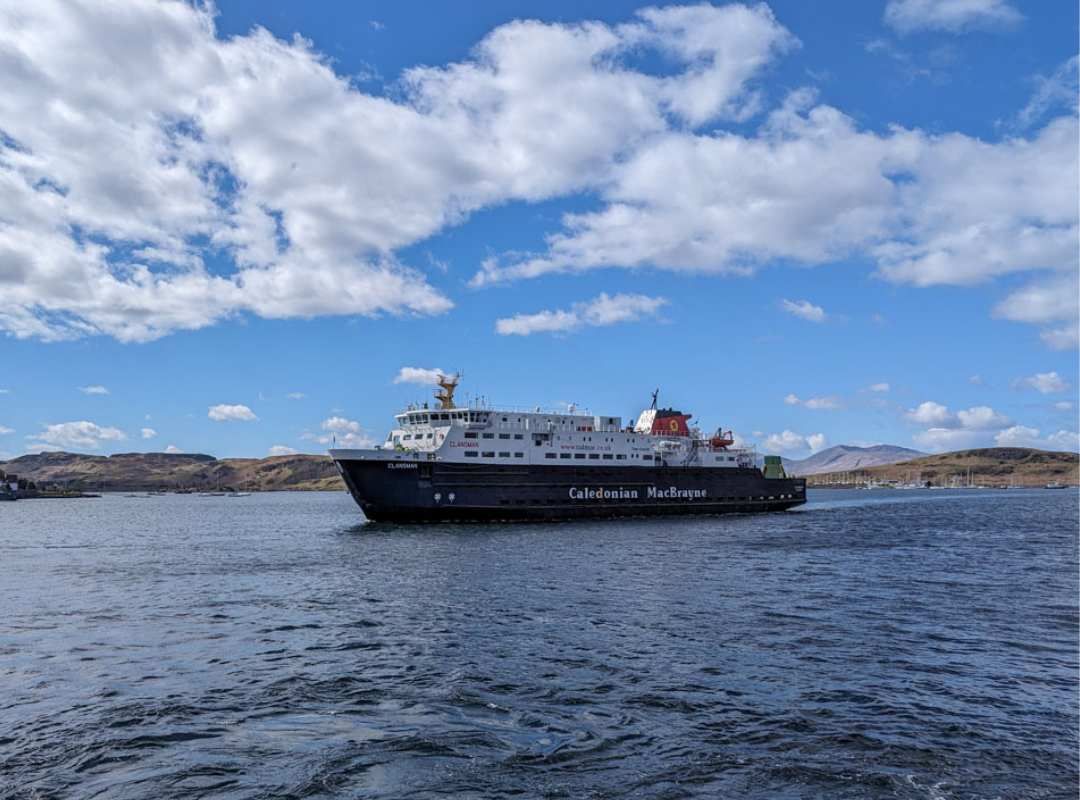A large ferry crosses a body of water on a blue but cloudy day. The hills of Mull can be seen in the distance in Scotland. 