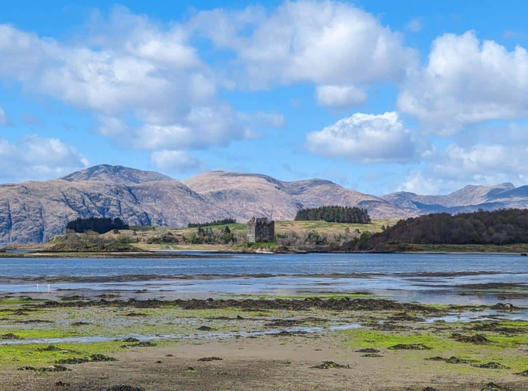 11 Best Day Trips From Glasgow By Public Transport 13 A shallow loch with the tide far out. Across it is an island with a fortress castle in the distance surrounded by mountains. Castle Stalker in Scotland.