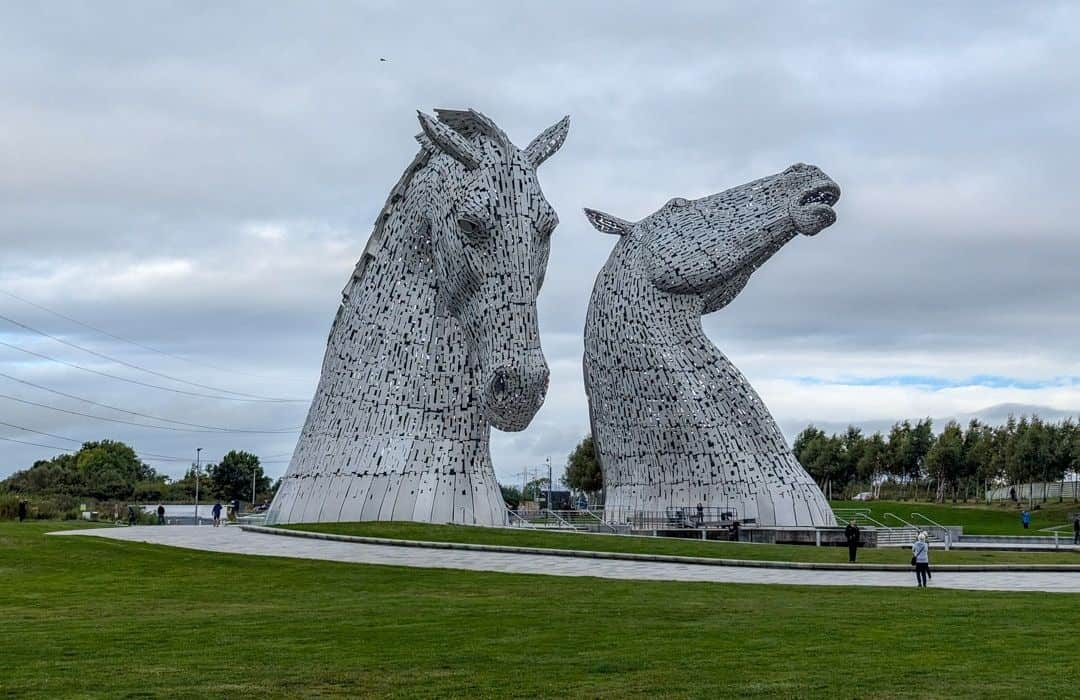11 Best Day Trips From Glasgow By Public Transport 7 Two silver metal horse heads rise up from manicured green grass ith people looking up at them. The Falkirk Kelpies in Scotland.