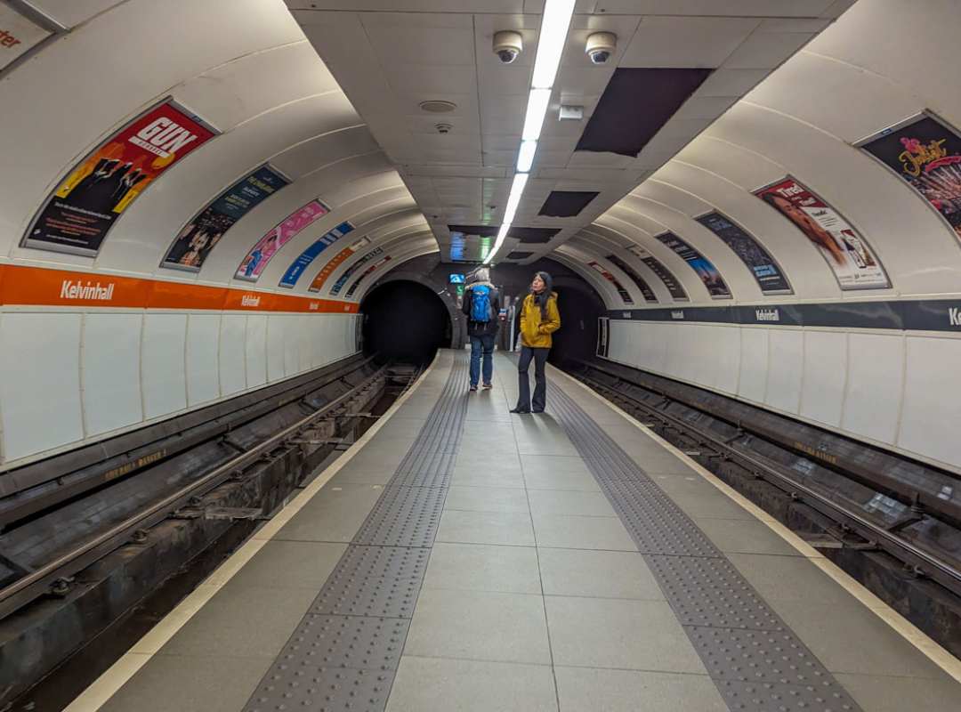 An undergroud tunnel with train tracks and posters on lit up white walls. Two people stand waiting on the small platform on the subway in Glasgow. 