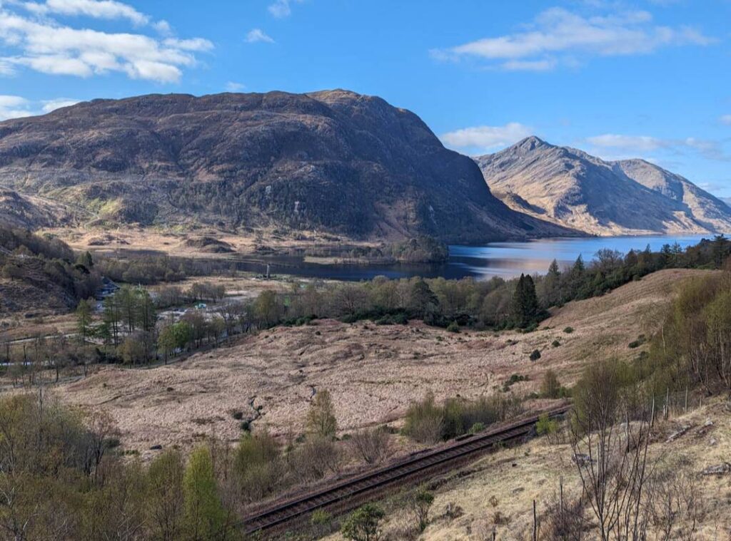 A craggy landscape with a railway line in the foreground and a blue loch surounded by mountains in the background. View from the Glenfinnan Trail. 