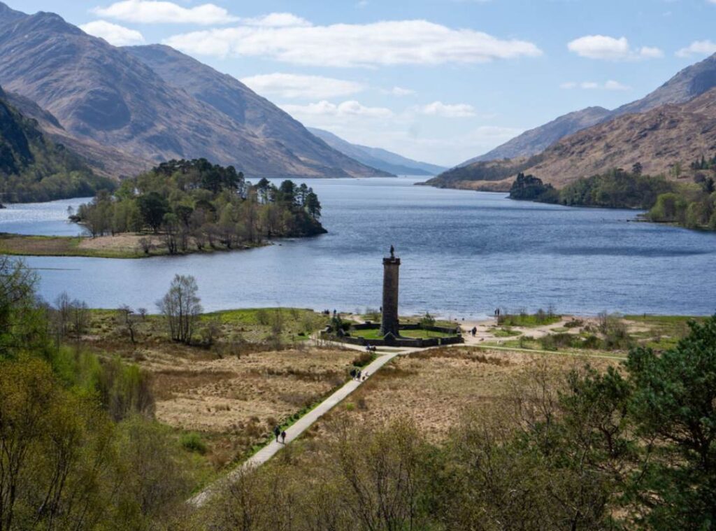 A tall tower stands with a statue on top beside a long lake. There is an island to the side with trees on it. Glenfinnan Monument. 