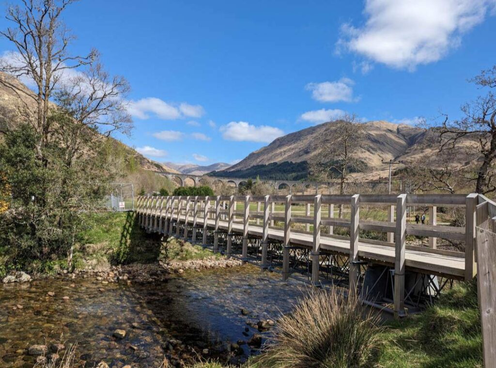 A wooden bridge crosses a shallow pebbly river. In the distance there are mountains anda glimpse of the top of the Glenfinnan Viaduct. 