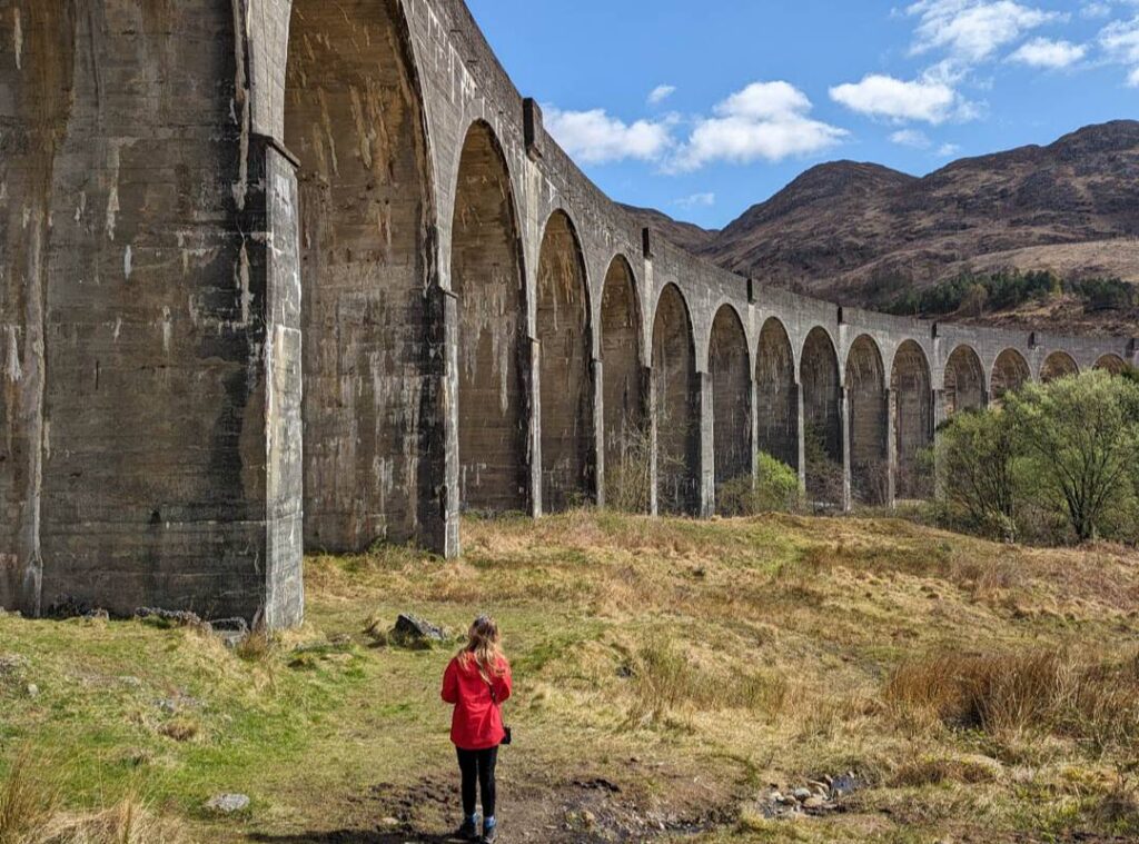 A girl in a red jacket stands facing the tall cement archways of Glenfinnan Viaduct with rugged mountains behind. 